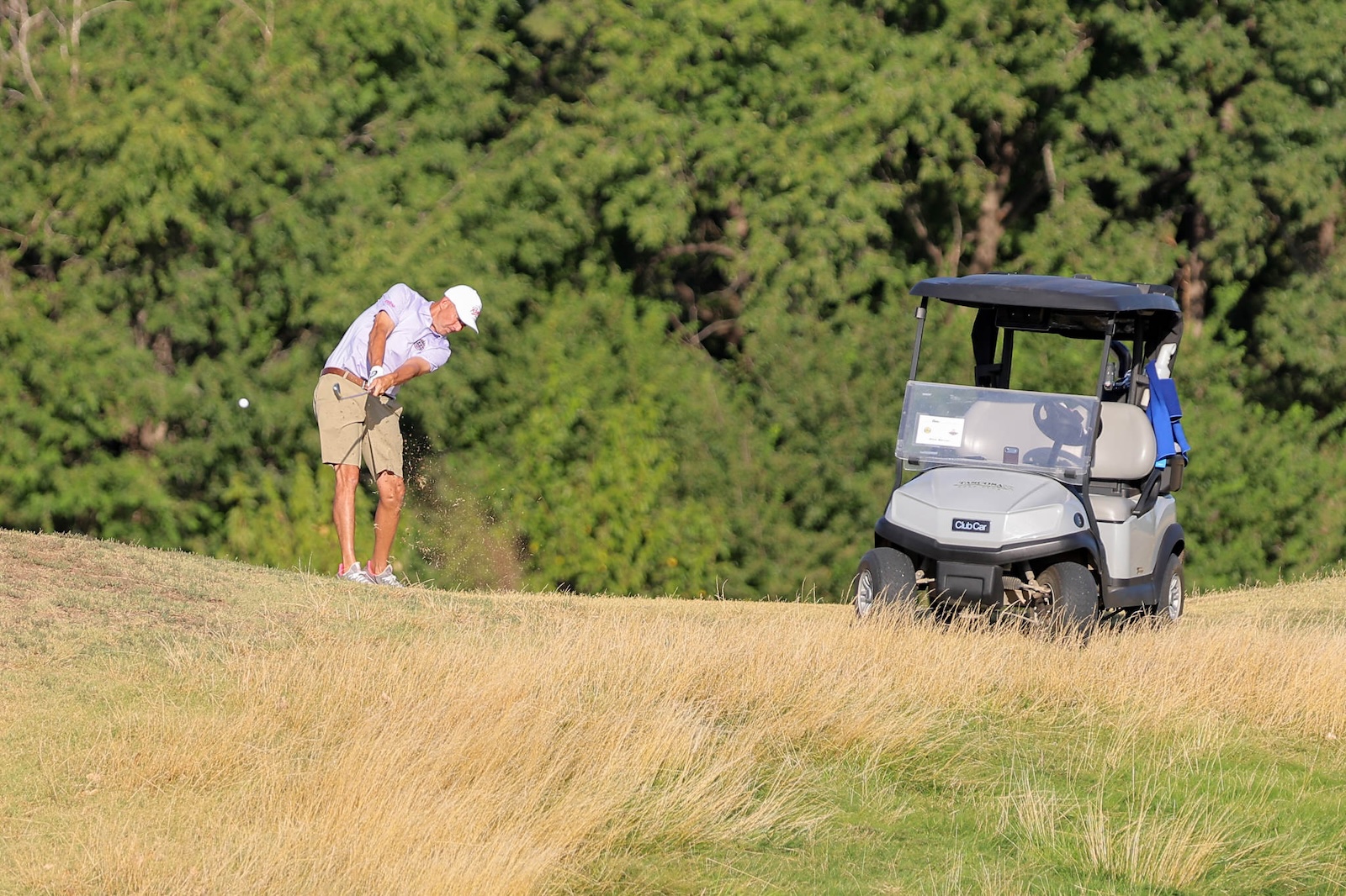 Male golfer in a white cap swings a club on a grassy hill beside a golf cart.