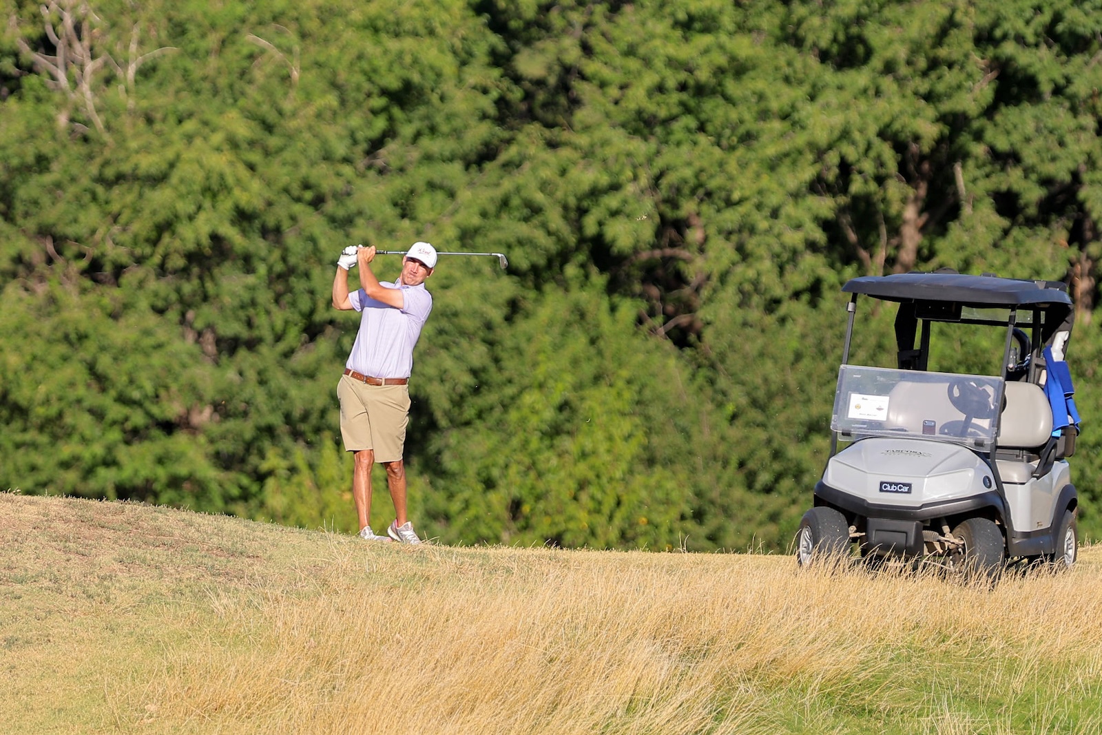 Golfer swinging a club on a sunny course beside a golf cart.