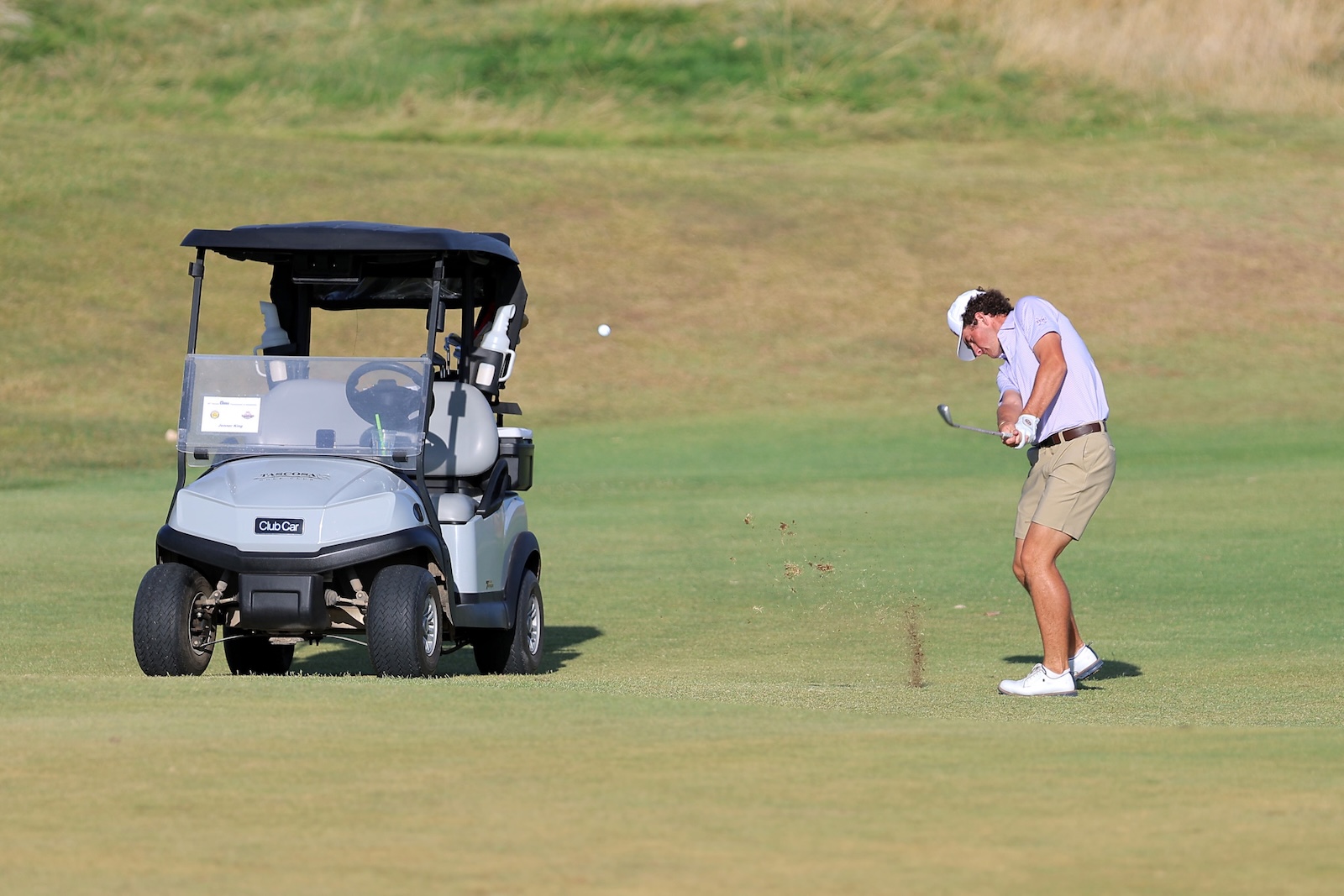 Golfer in a light shirt and shorts swings at a golf ball on a sunny course beside a parked gray golf cart.