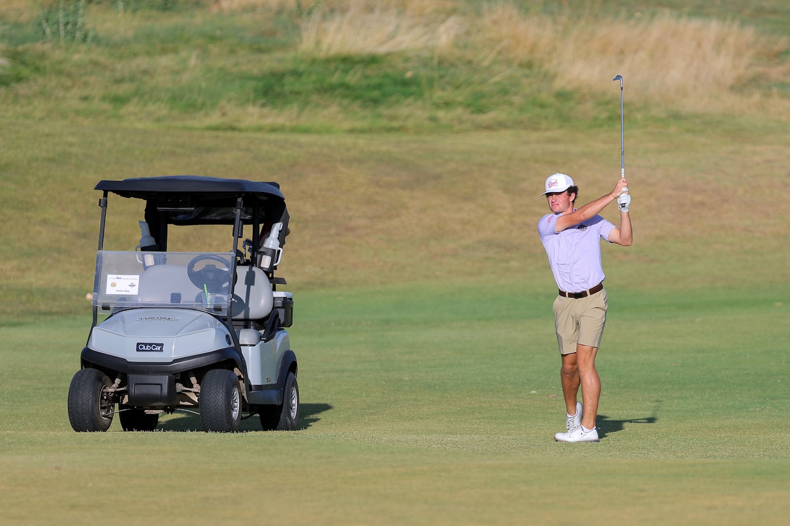 Golfer in a light purple shirt and beige shorts swings a club on a green fairway beside a gray golf cart.