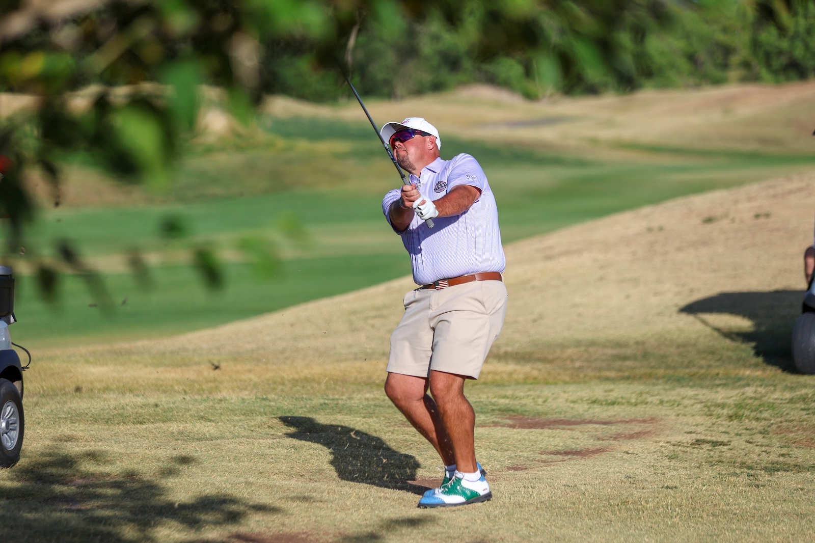 Golfer mid-swing on a sunny golf course: light purple polo, beige shorts, white cap, sunglasses, and green-and-white shoes. A golf cart is visible on the left.
