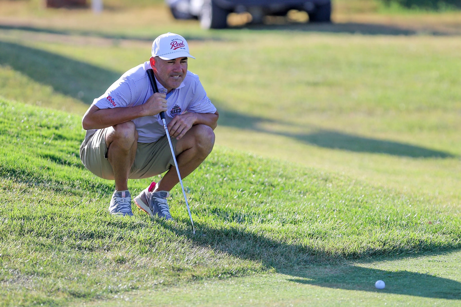 Man in a white cap and striped polo crouches on a grassy golf course, holding a putter beside a golf ball.