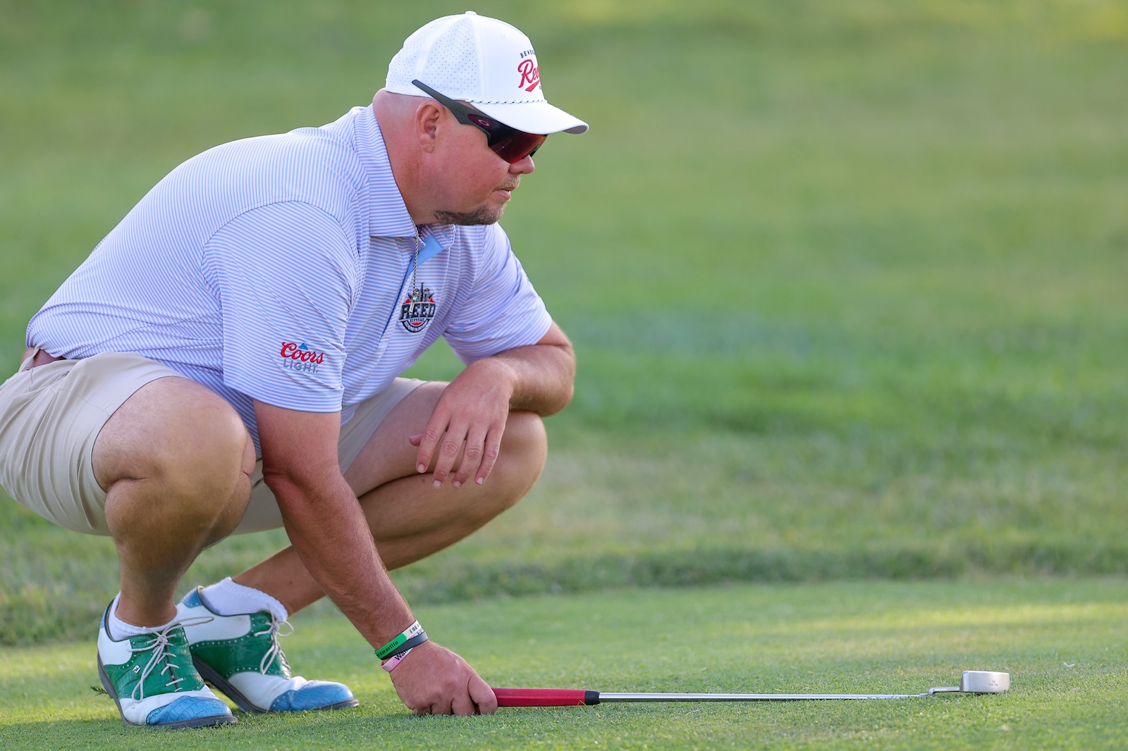 Crouching golfer lines up a green putt with a red putter, wearing a white cap, sunglasses, and a striped polo on the course.
