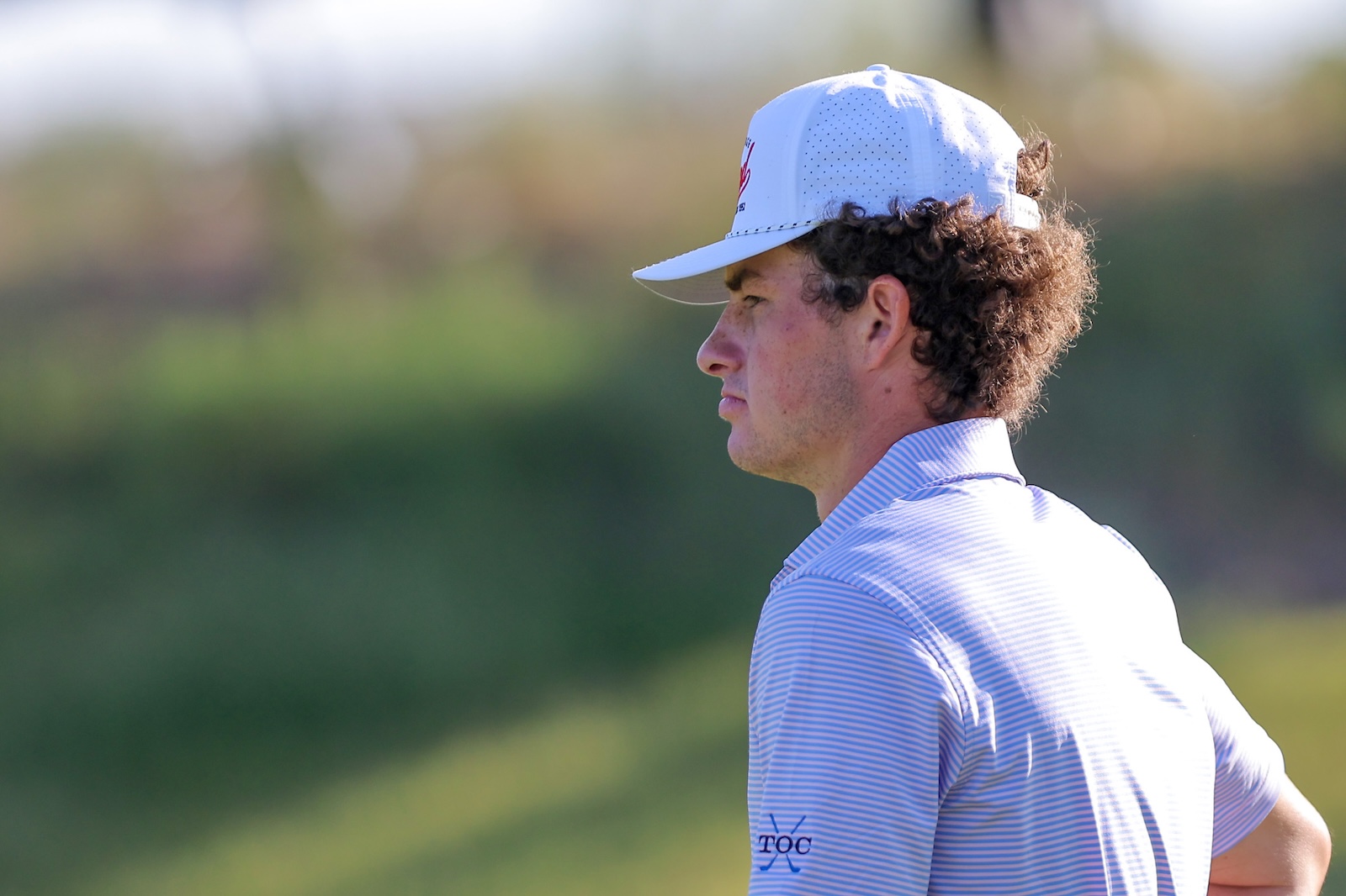 Side profile of a man wearing a white cap and light blue striped polo, looking to the left on a sunny golf course.