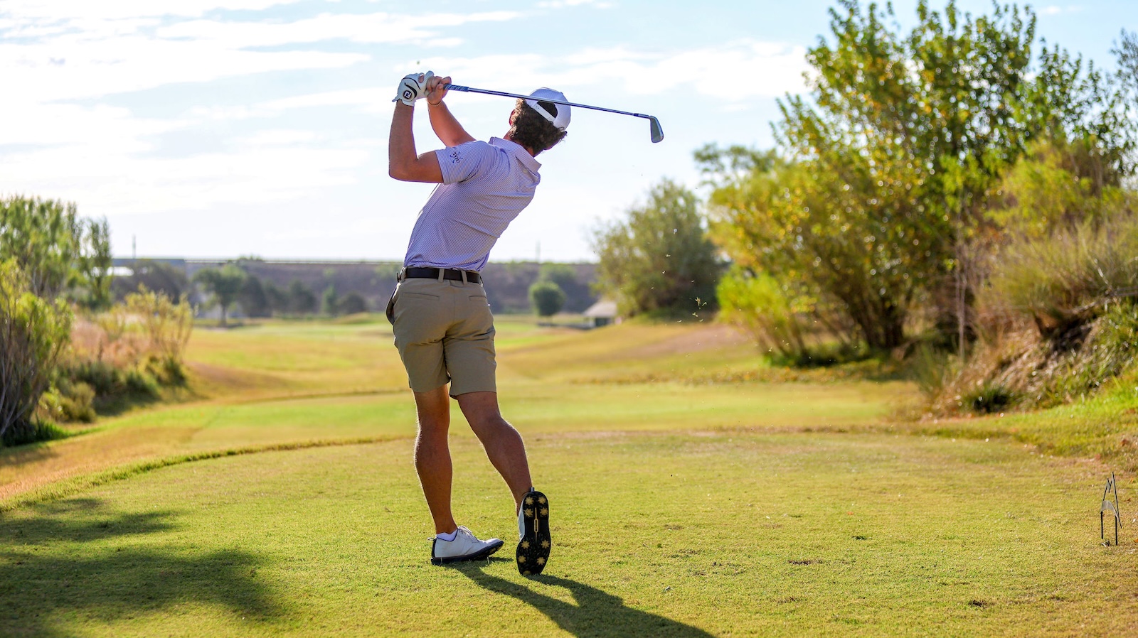 Male golfer mid-swing on a sunlit golf course, driving from the tee with a driver smacked overhead of the backswing