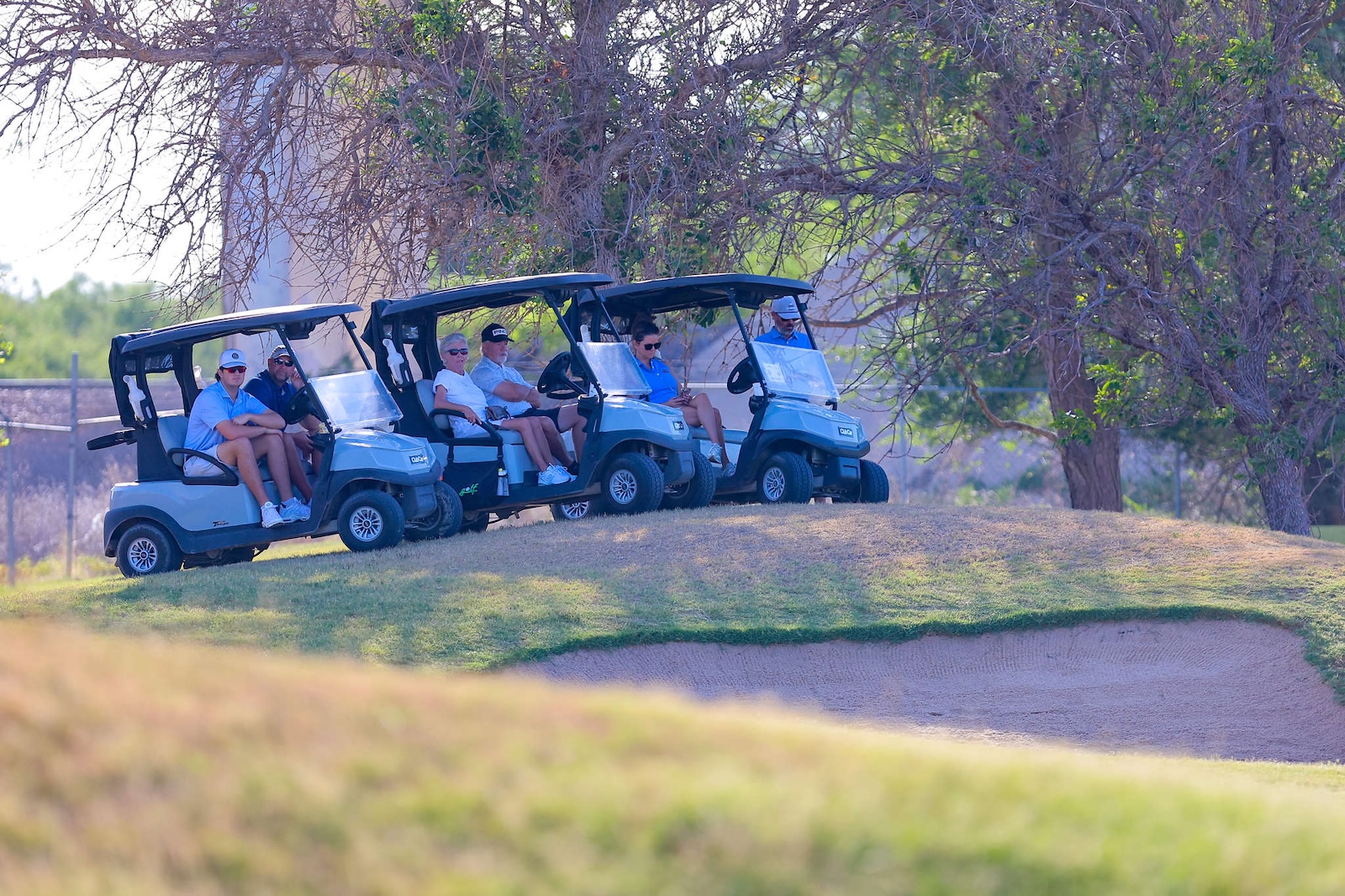 Six people seated in four blue golf carts lined up on a grassy hill at a golf course.