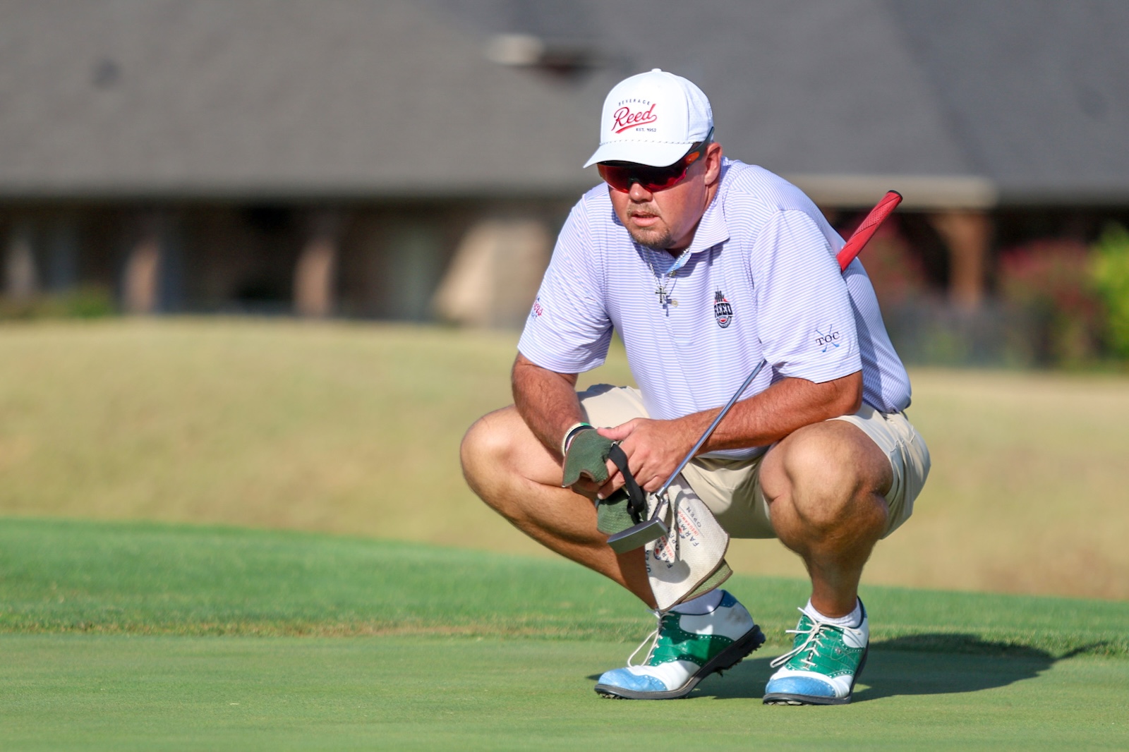 Golfer crouching on a putting green, gripping a putter with a glove, wearing a white cap and sunglasses.