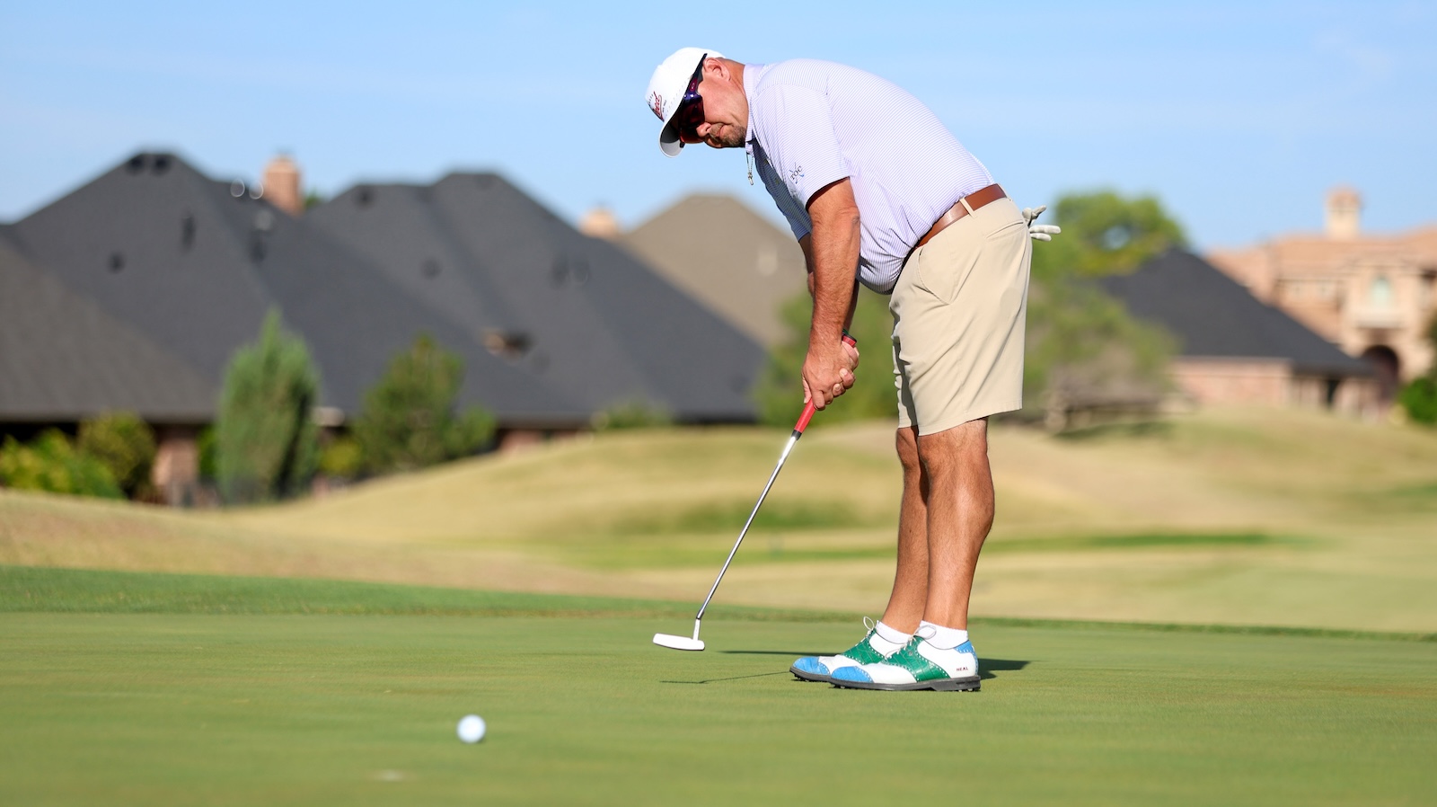 Golfer preparing to putt on a sunny golf course green, wearing a white cap, striped polo, beige shorts and teal sneakers, with a ball in the foreground.
