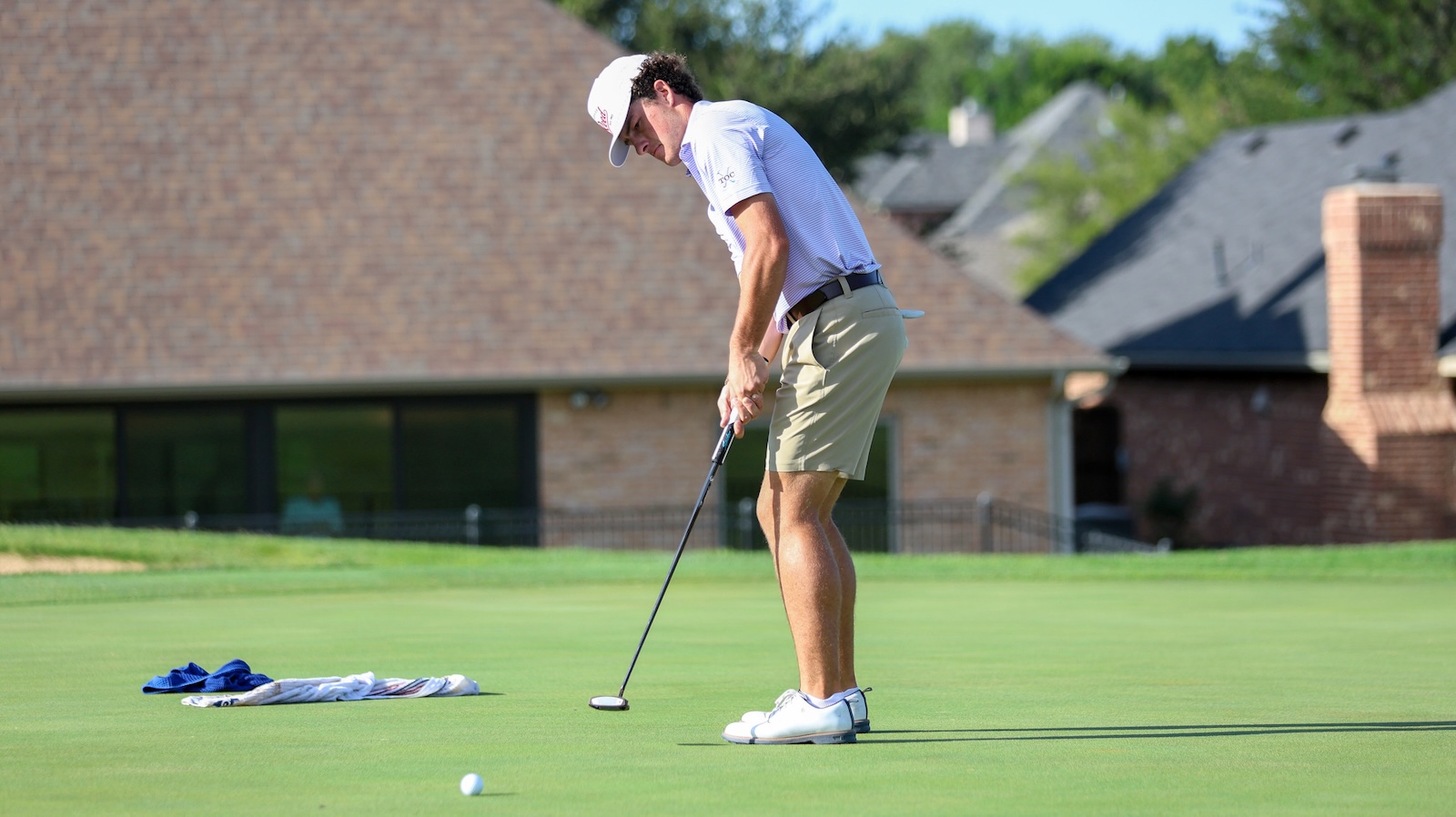 Golfer lining up a putt on a well-manicured green, wearing a white cap and khaki shorts.
