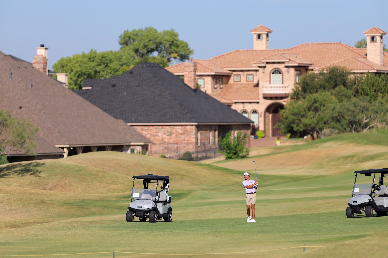 Golfer in light shirt and beige shorts swings on a green fairway with two golf carts nearby and large brick houses in the background.