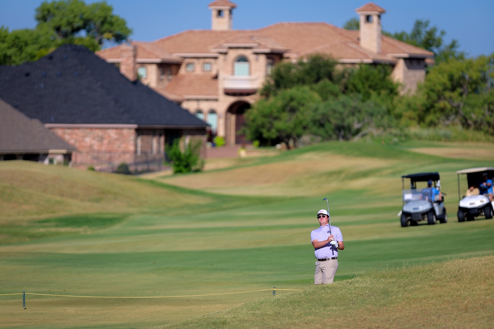 A golfer in a light shirt and cap swings a club on a sunny golf course, with carts and a mansion in the background.