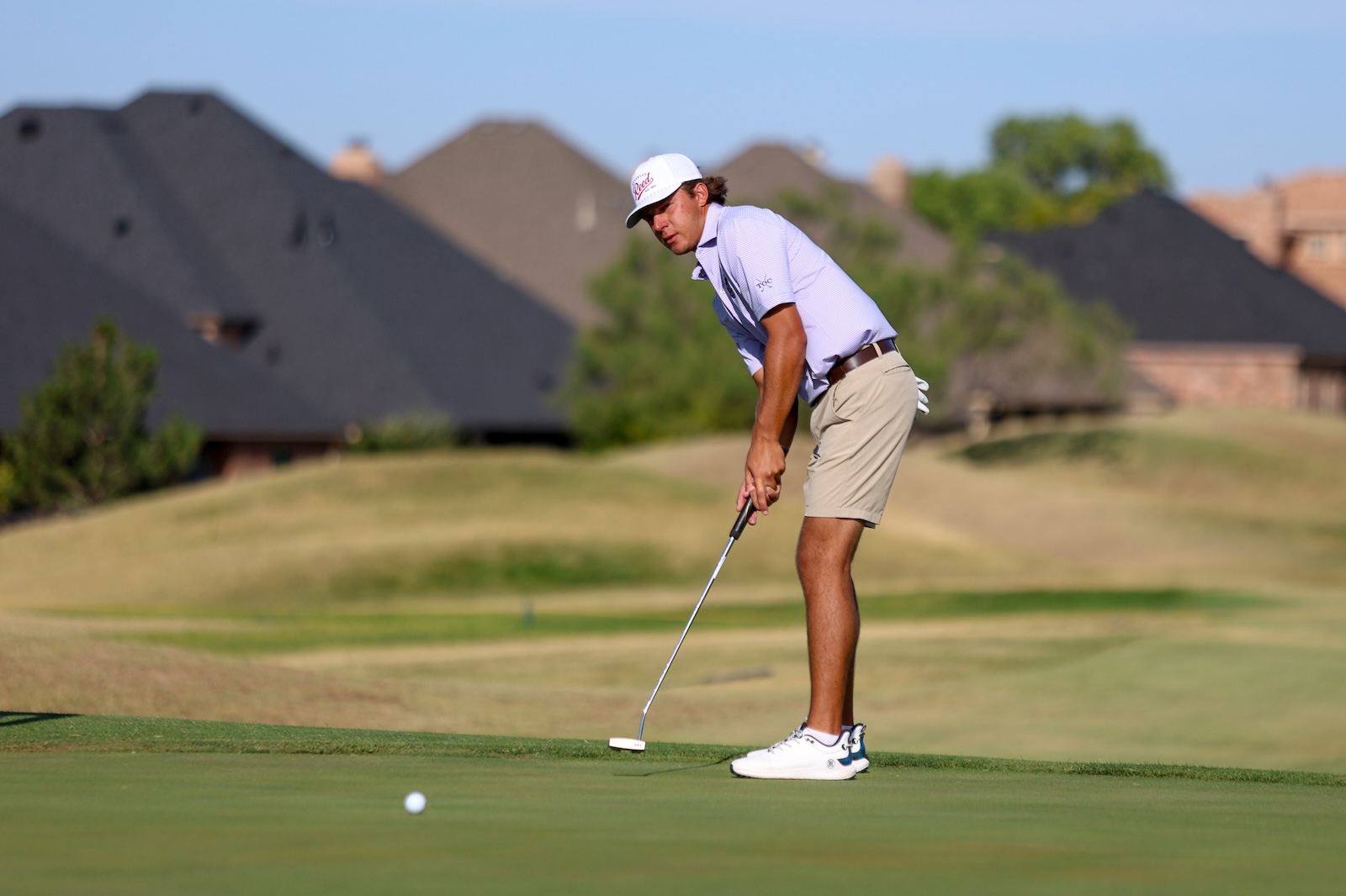 Golfer in a white cap and lavender shirt prepares a putt on a green with suburban homes in the background.