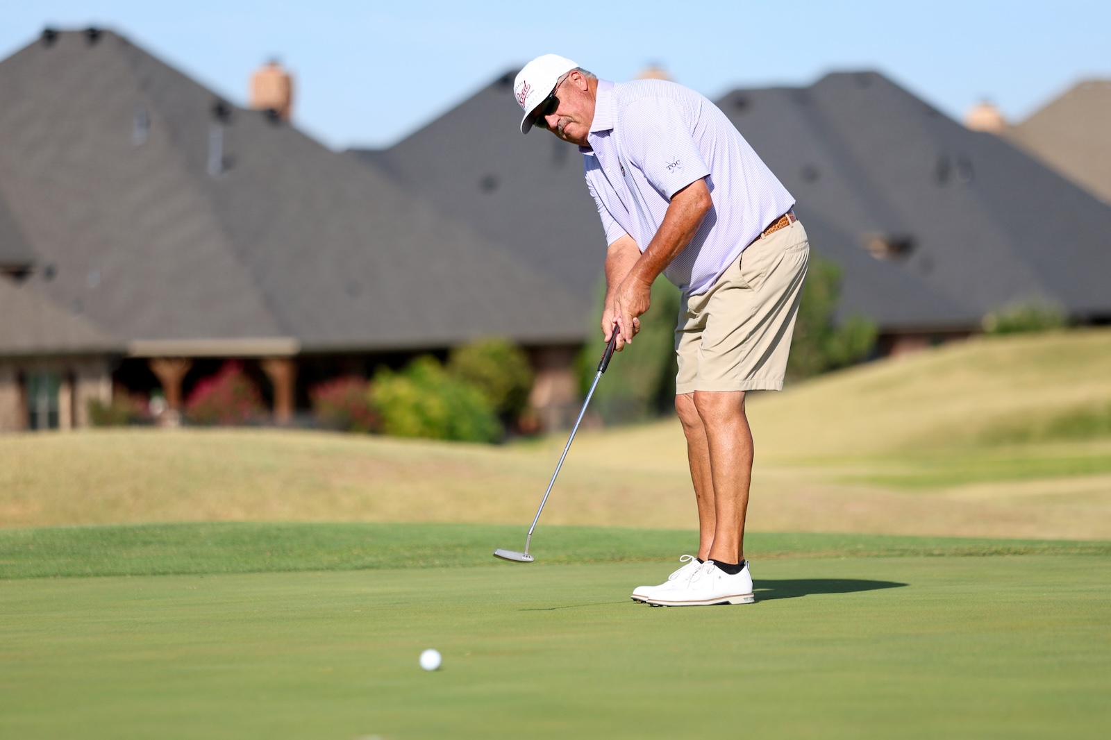 An older man putting on a golf green, wearing a light polo, shorts, white cap and sunglasses with houses in the background