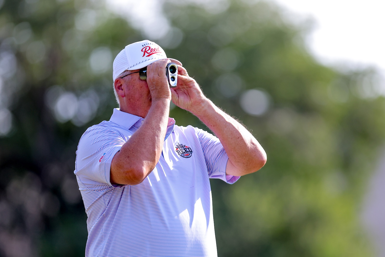 Man in a light purple striped polo shirt and white cap looking through small binoculars outdoors