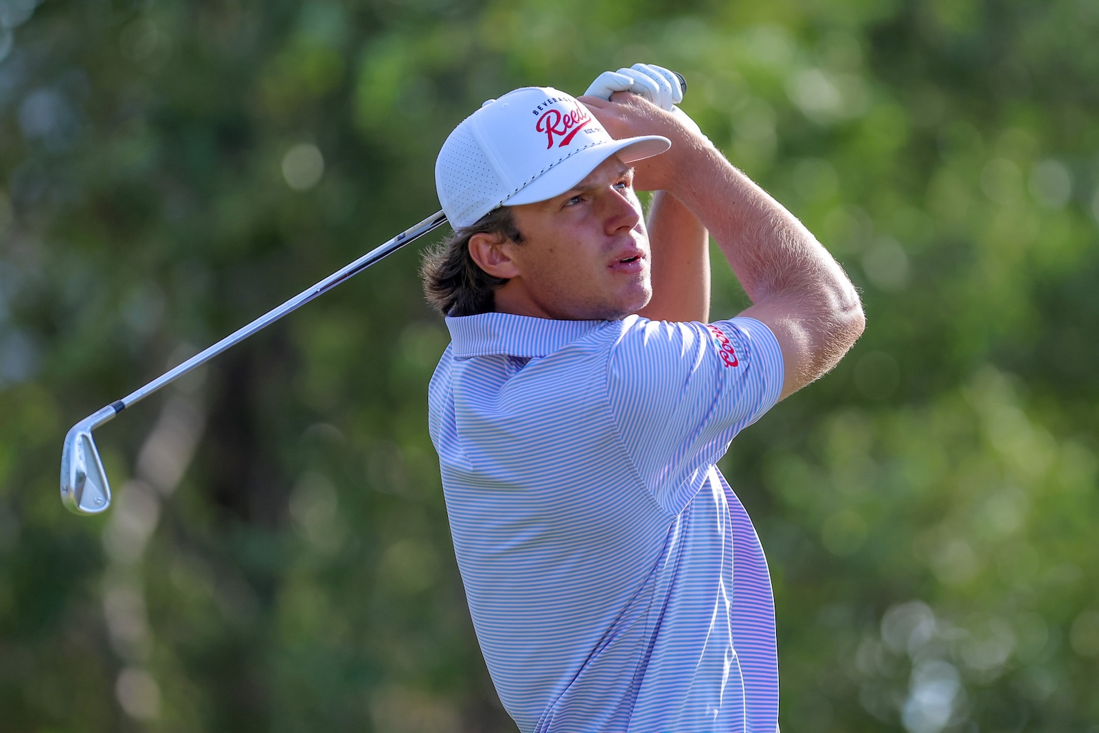 Golfer in a blue striped polo and white cap mid-swing on a sunny green course, focusing on the shot.