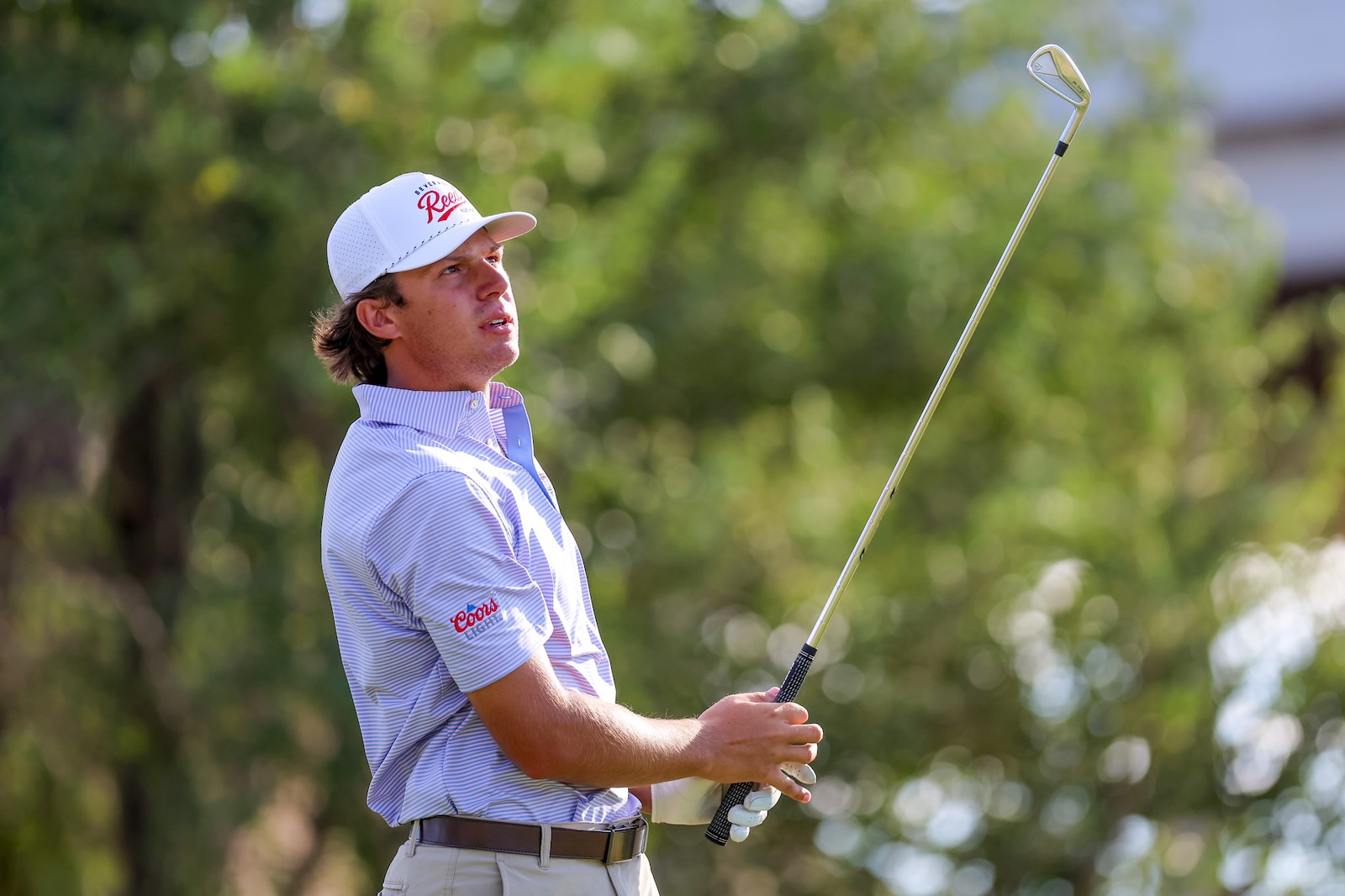 Man in a striped polo and white cap holding a golf club mid-swing outdoors.