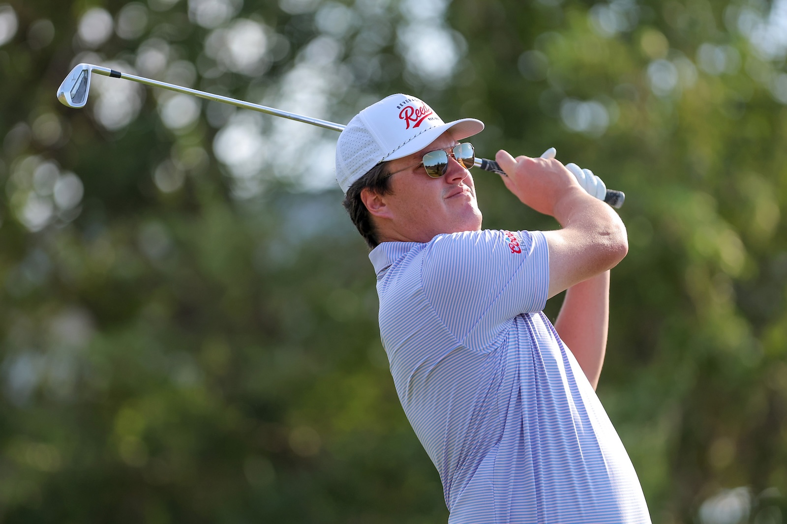 Man in a striped polo and white cap swings a golf club outdoors, wearing sunglasses.