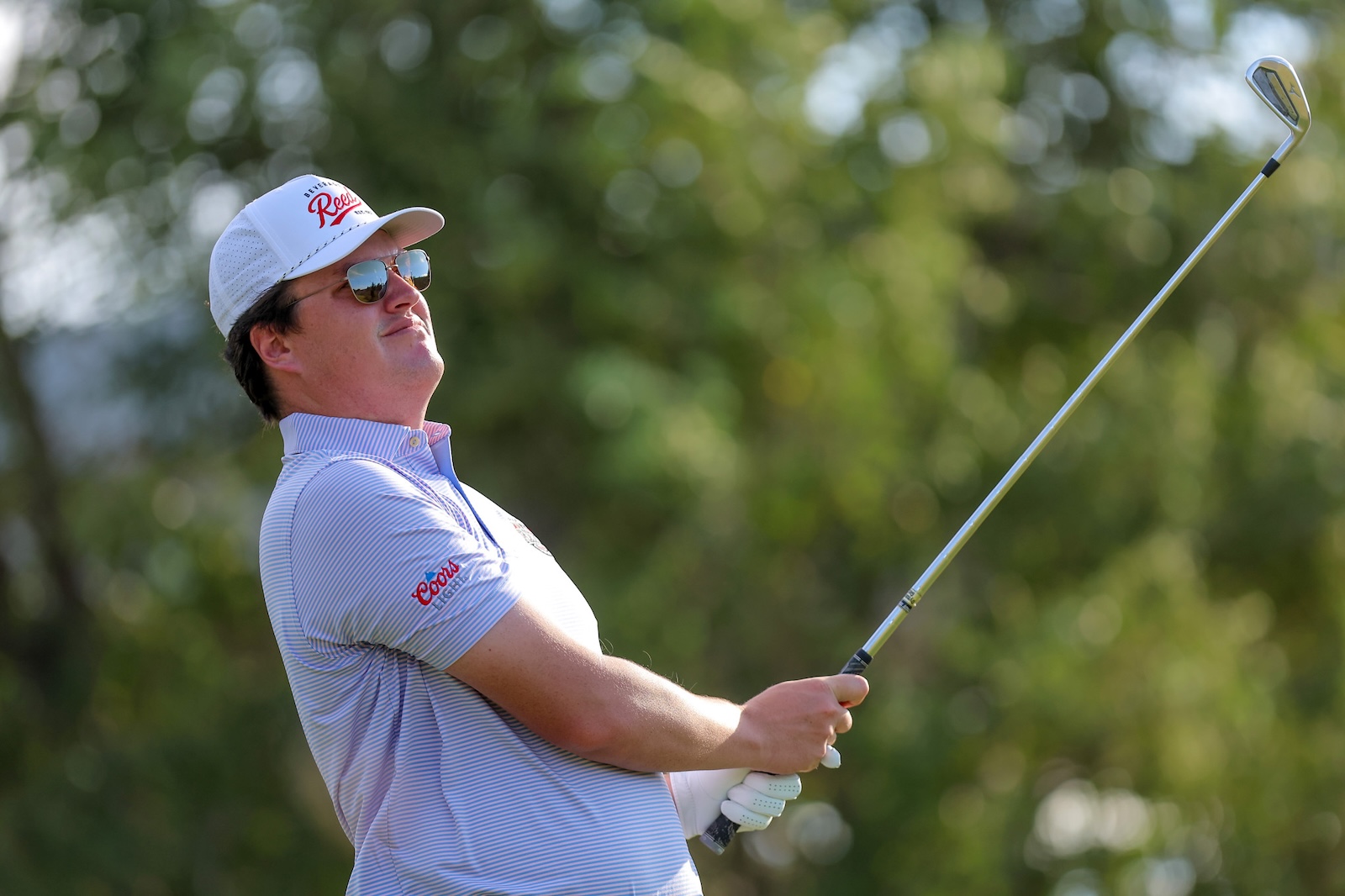 Male golfer in a striped polo and white cap swings a golf club on a sunny course, wearing sunglasses against a blurred green background.