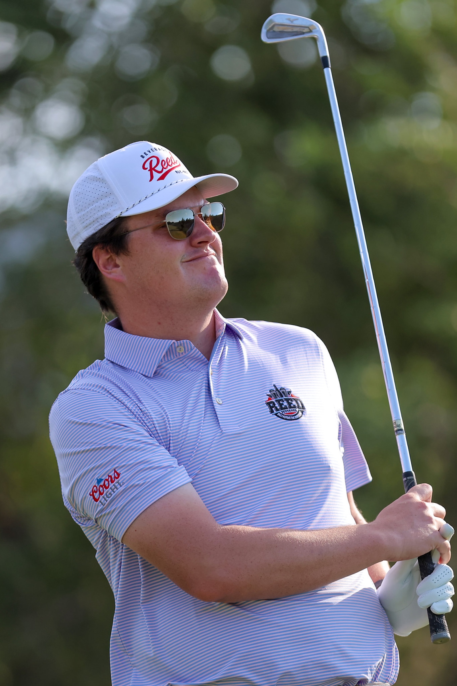 Man wearing a white cap and sunglasses, in a light-striped polo, holding a golf club on a sunny day.