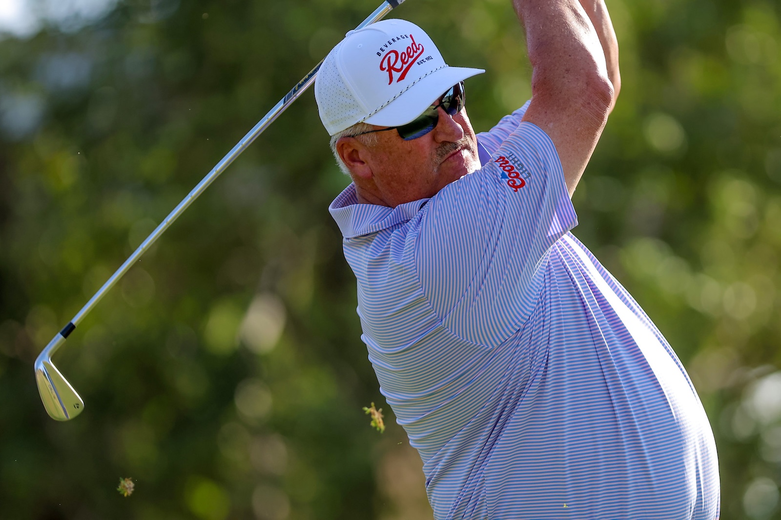 Male golfer in a white cap and sunglasses mid-swing on a sunny golf course