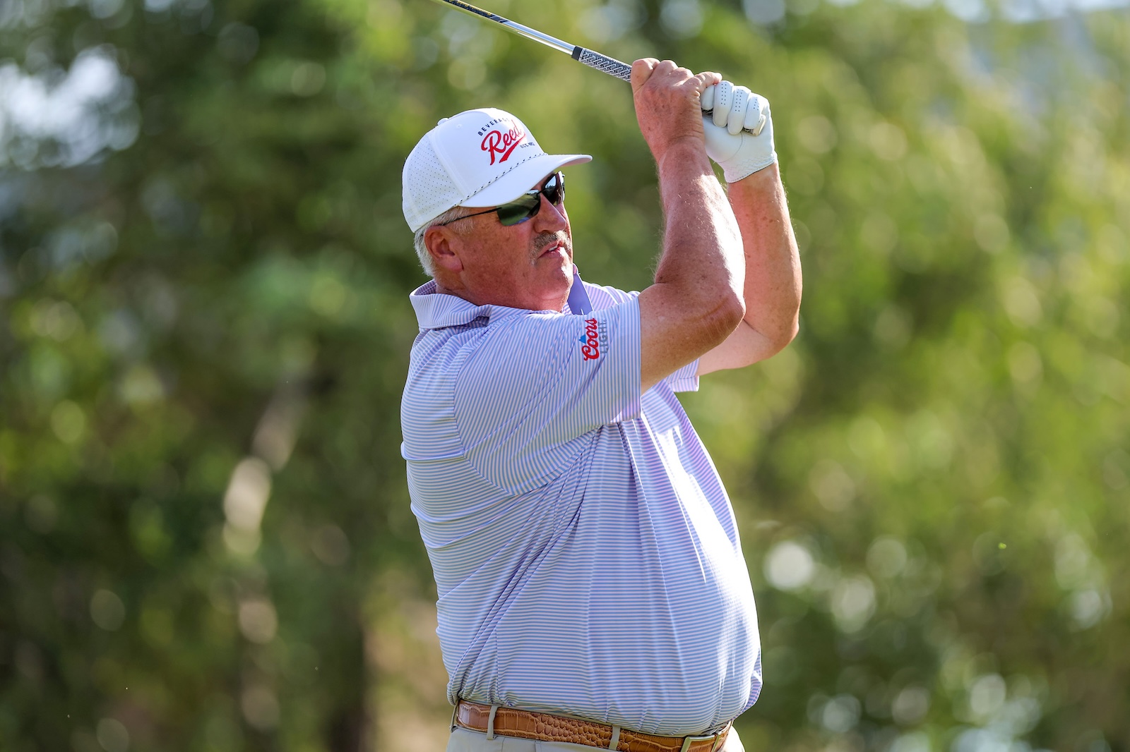 Older man golfer in a white cap, sunglasses, and striped polo swinging a club outdoors.