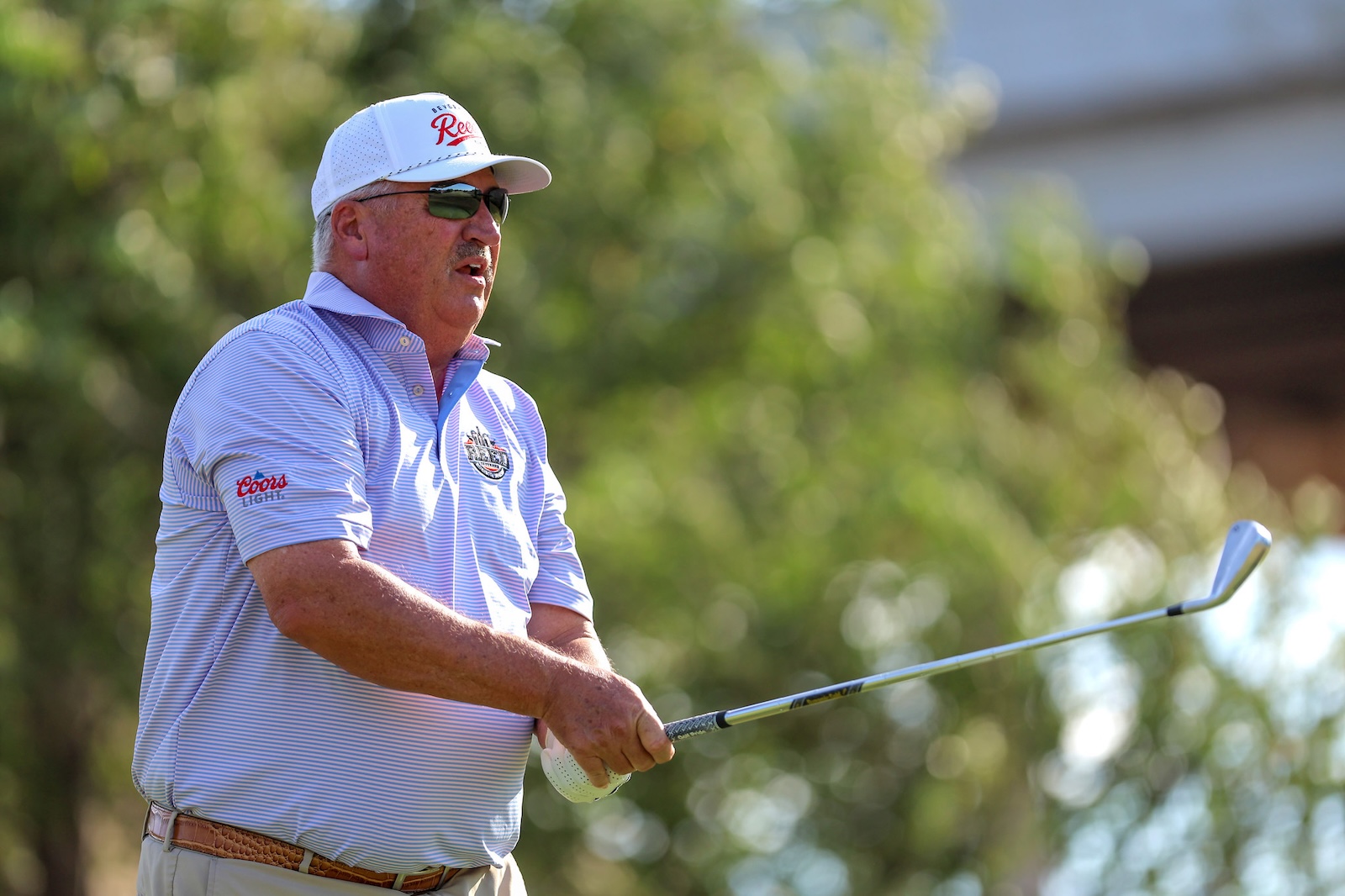 Older man wearing a white cap and striped polo swinging a golf club on a sunny day on the course
