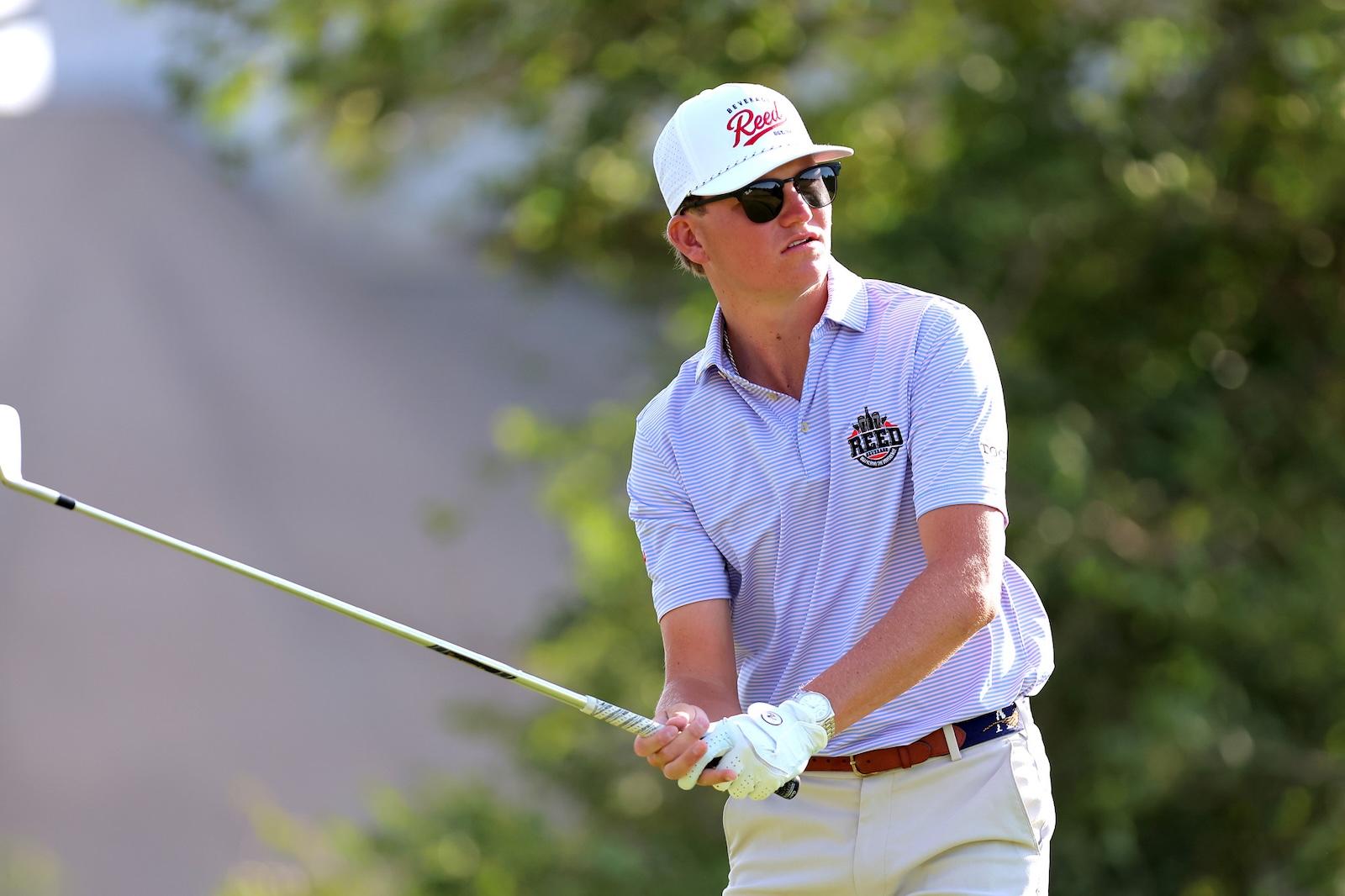 Male golfer in a striped polo and white cap mid-swing on a sunny golf course, wearing sunglasses and a glove on his left hand.
