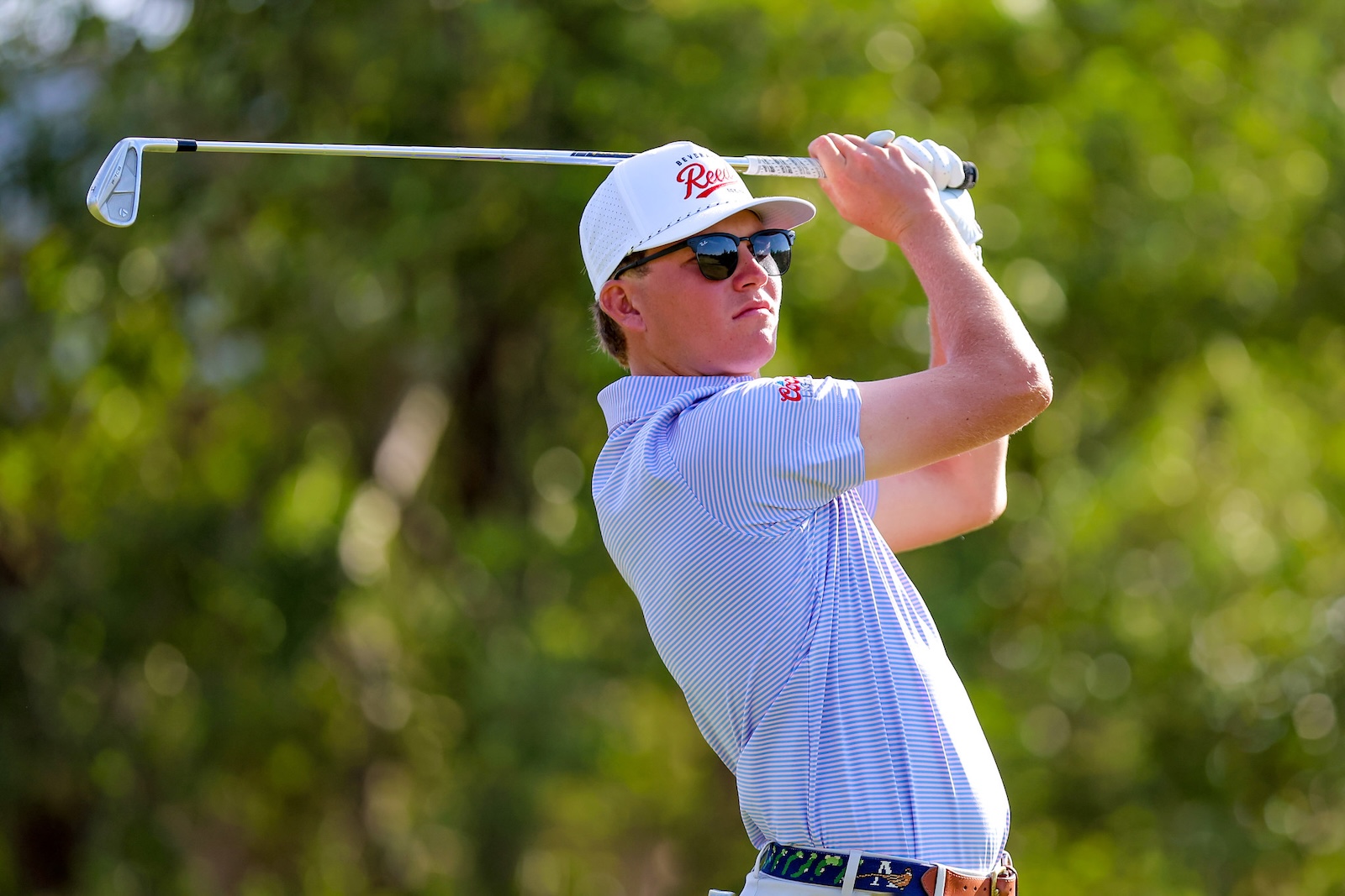 Male golfer in sunglasses swinging a club overhead on a sunny golf course, wearing a white cap and light blue striped polo.