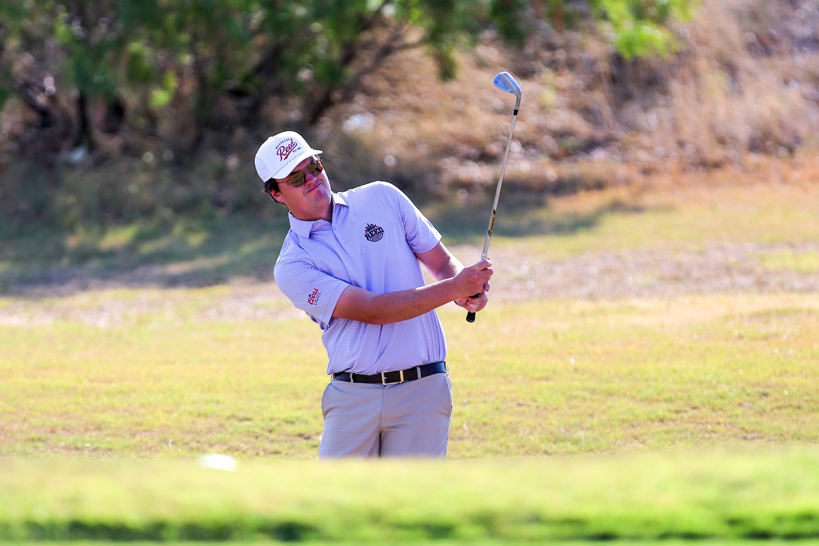 Male golfer in a light blue polo and white cap mid-swing on a sunny golf course