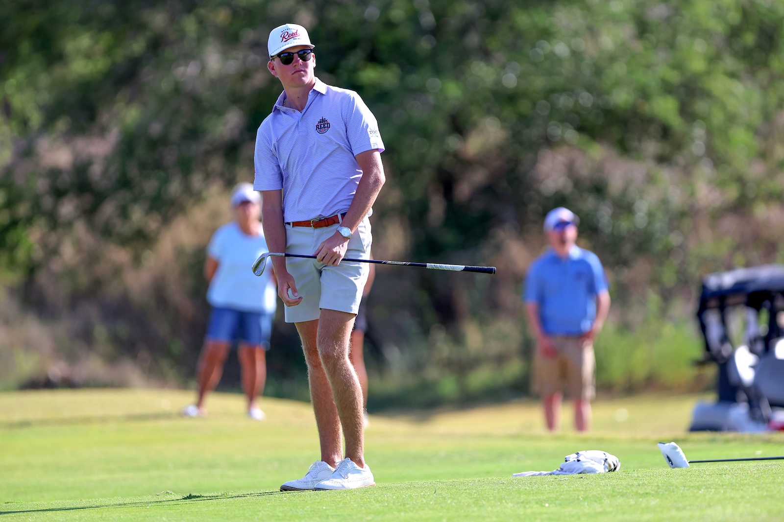 A male golfer in a light purple polo and white cap prepares to swing on a greenside area, holding a putter-like club.
