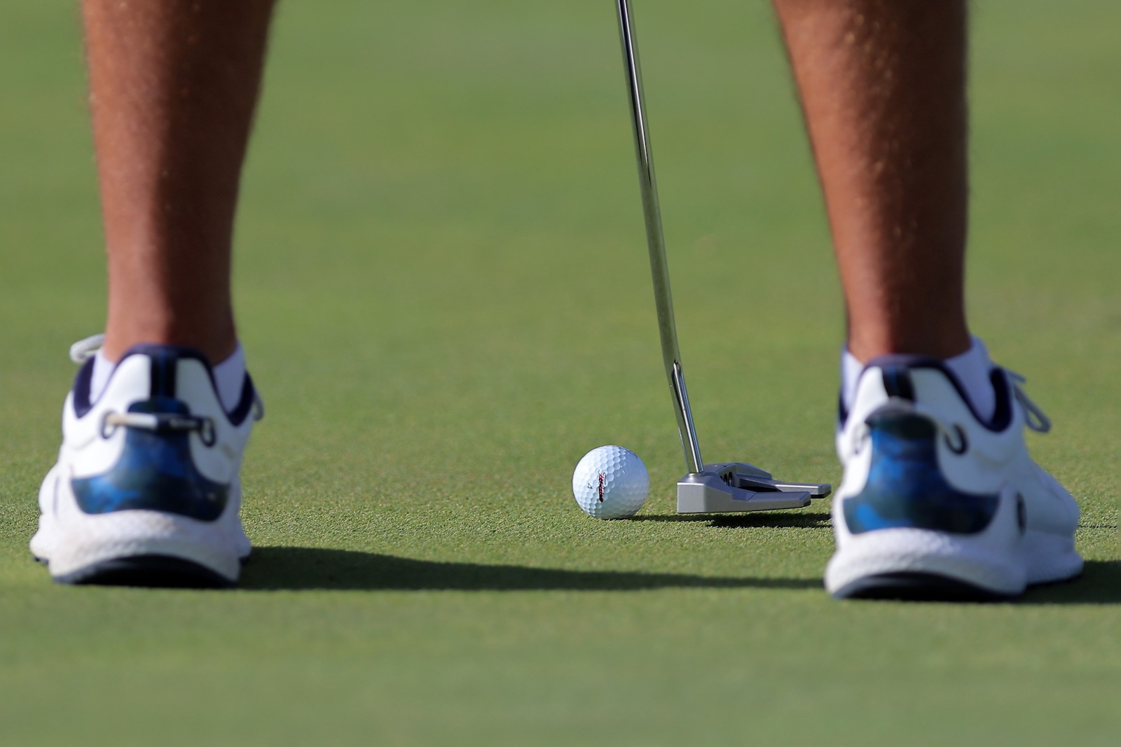 Golfer's feet in white and blue shoes as a putter aims at a golf ball on the green.