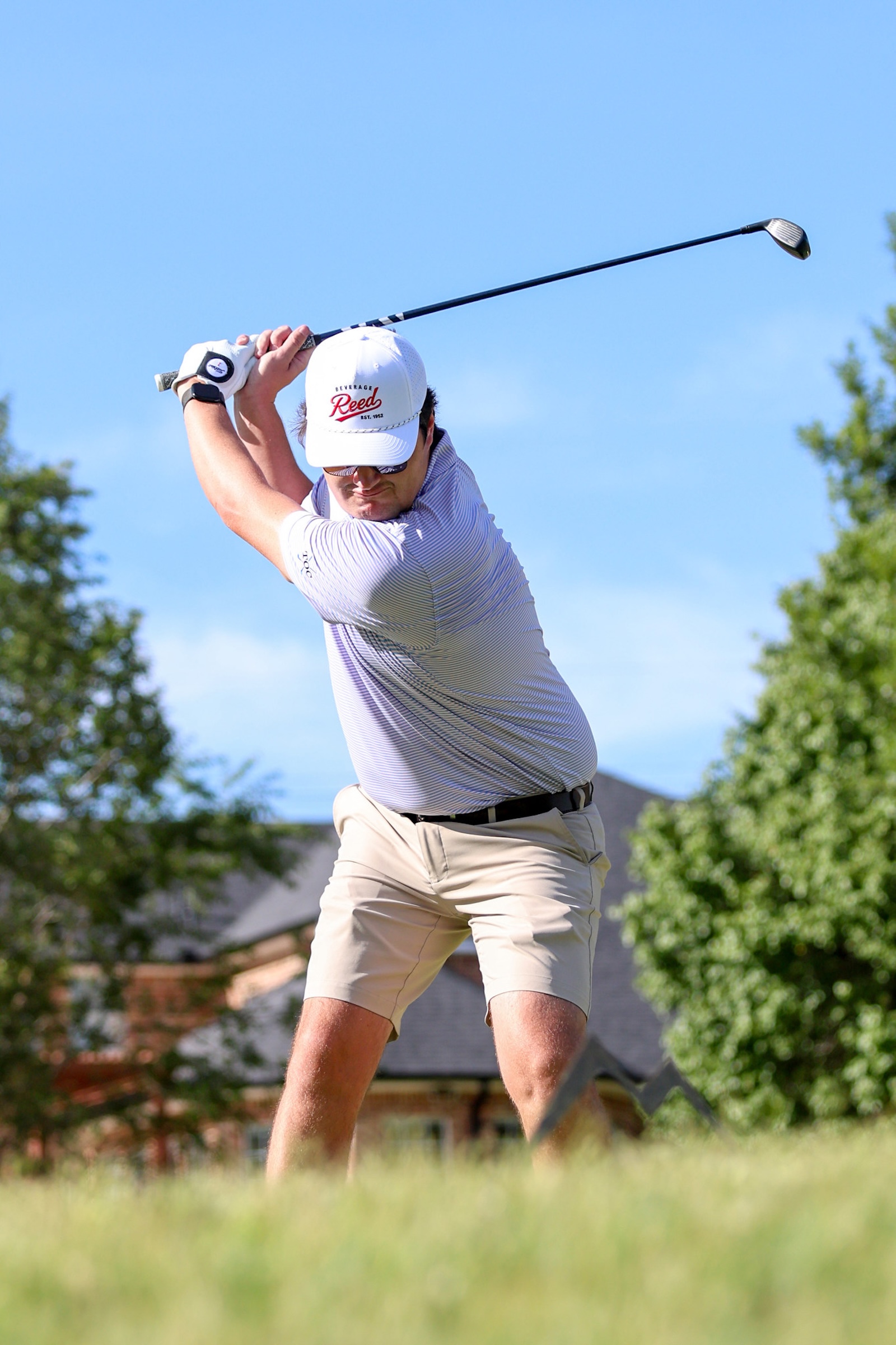 Male golfer in a white cap and striped polo mid-swing with a driver on a sunny day; suburban houses and green trees in background.