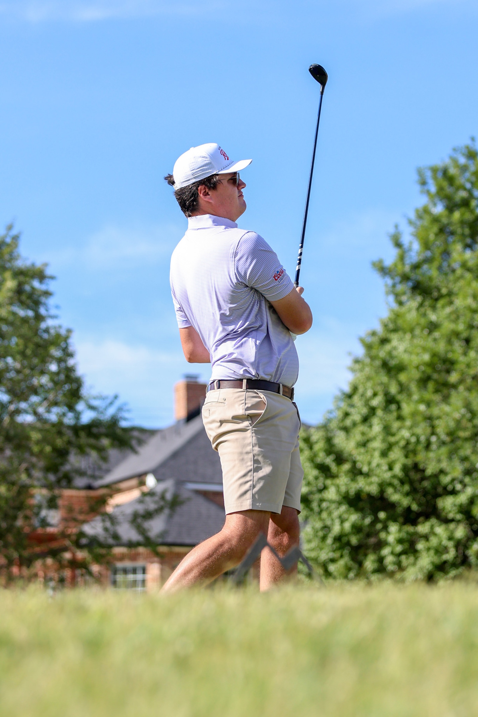 Male golfer in a white cap and striped polo swings a driver on a sunny day.