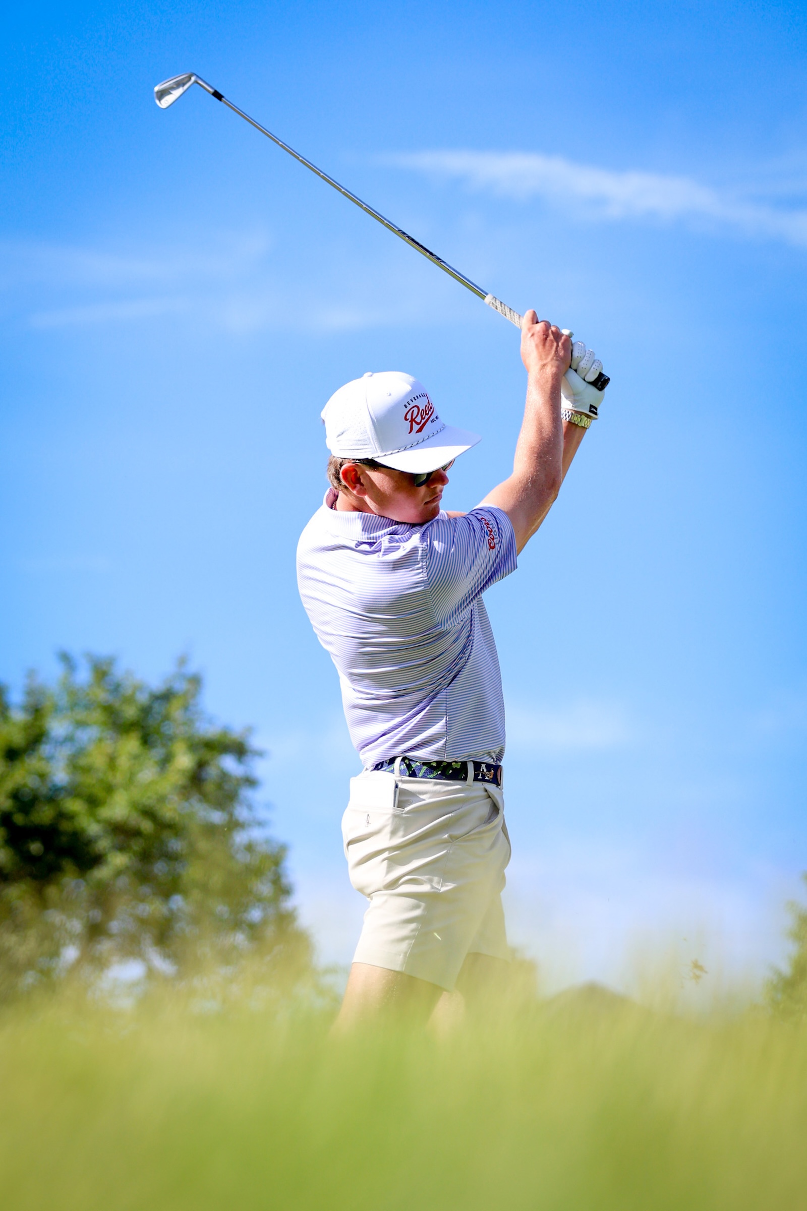 Golfer in a white cap and striped polo mid-swing against a clear blue sky