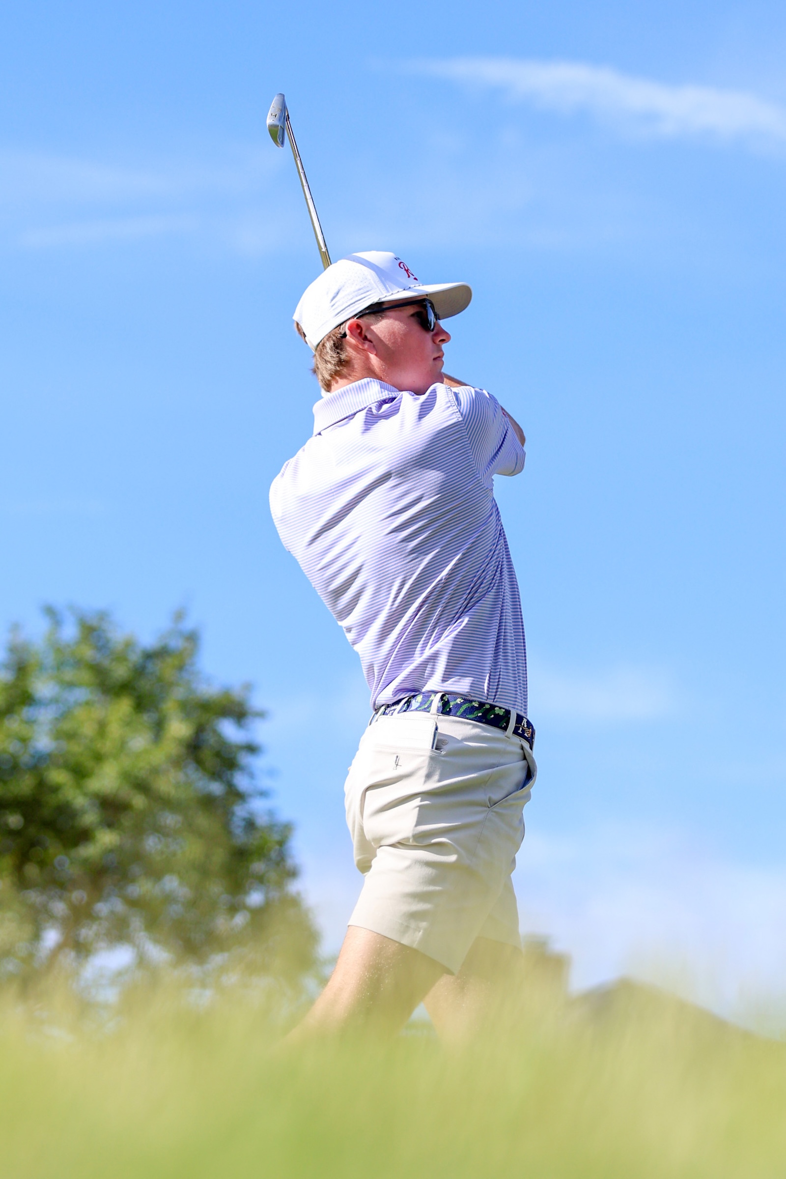 Golfer in a light striped shirt and white cap swinging a golf club against a bright blue sky.