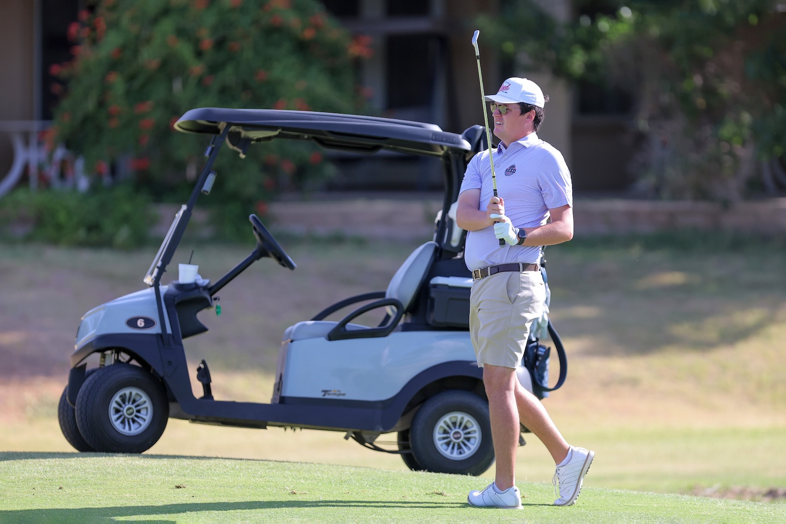 Man in a polo shirt and cap swings a golf club beside a blue golf cart on a sunny course.