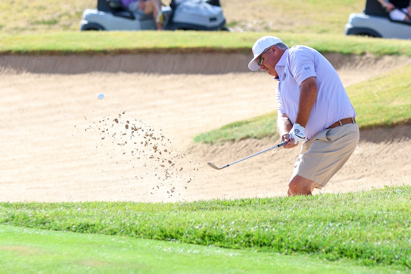 Golfer in a light polo and beige shorts hitting from a sand bunker as sand sprays into the air and a white golf ball rises nearby.