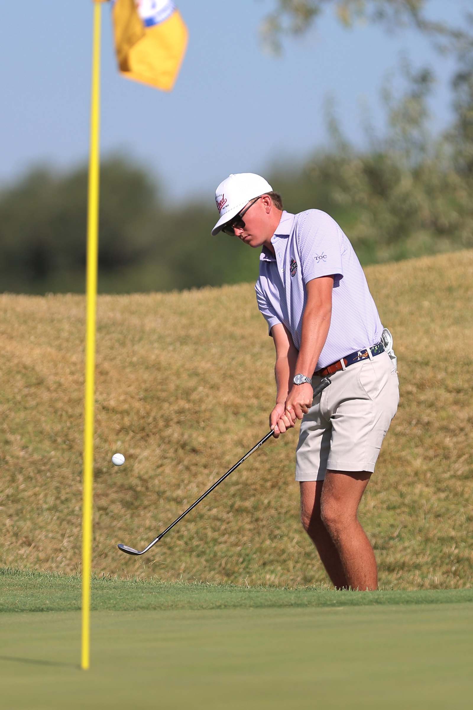 Male golfer preparing to putt on the green beside a yellow flagstick, wearing a white cap and striped polo shirt.
