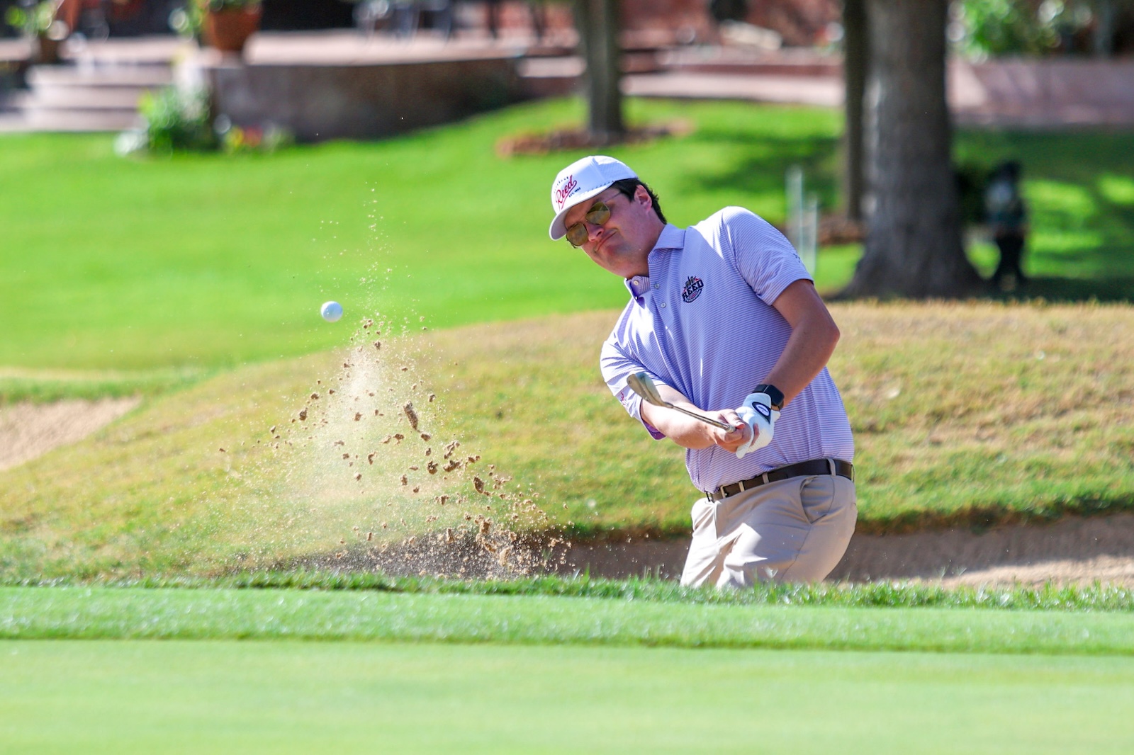 Golfer in a light purple polo and white cap hits a bunker shot; sand sprays as the ball lifts from the sand trap.