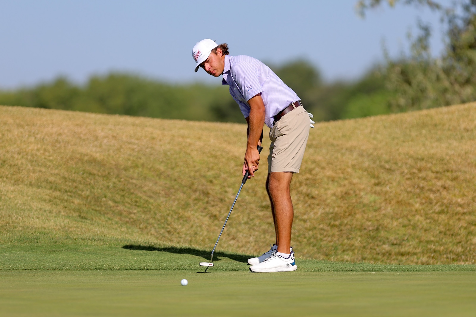 Golfer in a light purple shirt and beige shorts lining up a putt on the green, with a white ball nearby.