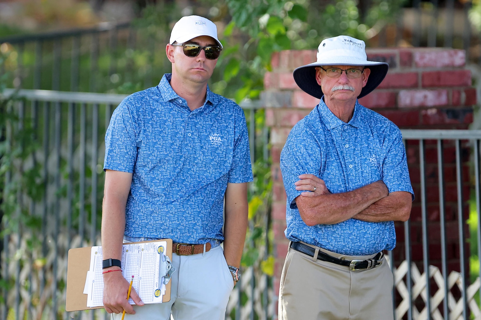 Two men in blue patterned polo shirts stand outdoors, one holding a clipboard and pencil, both wearing hats and sunglasses.