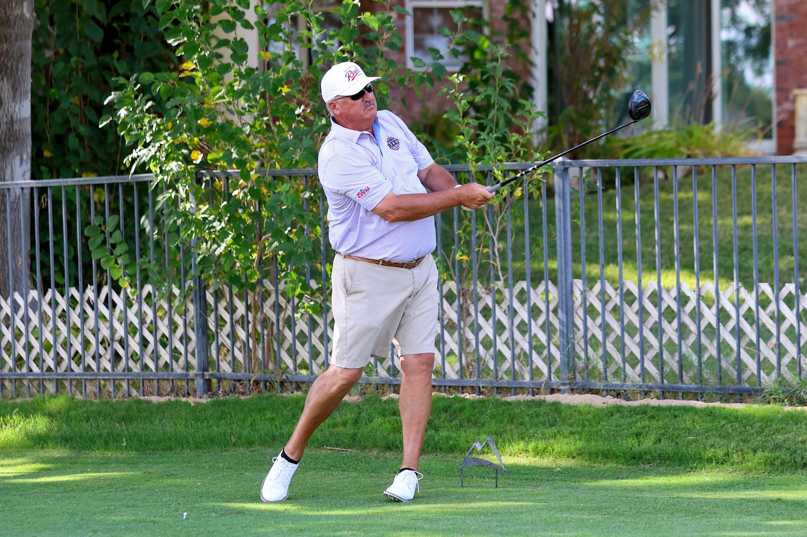 Man in a light polo and white cap swinging a golf club in a backyard with a metal fence and green grass.