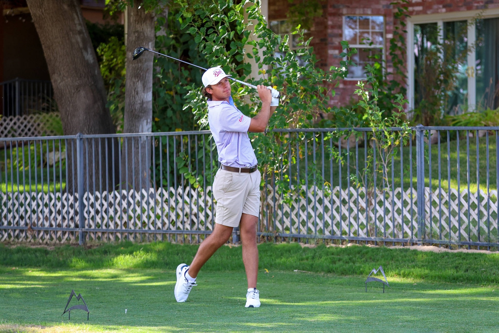 A man in a white cap and polo swings a golf club on a grassy backyard course behind a metal fence.