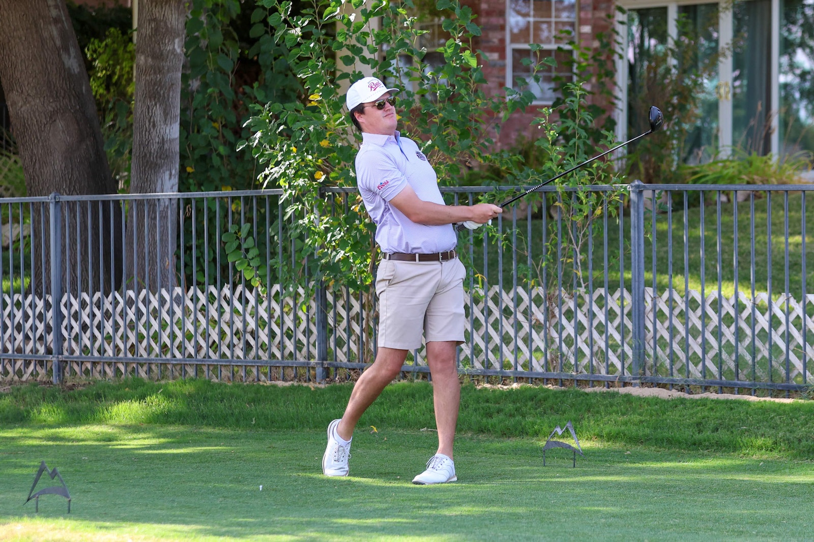 Man in a light purple polo shirt, beige shorts, and white cap swinging a golf club on a grassy backyard.
