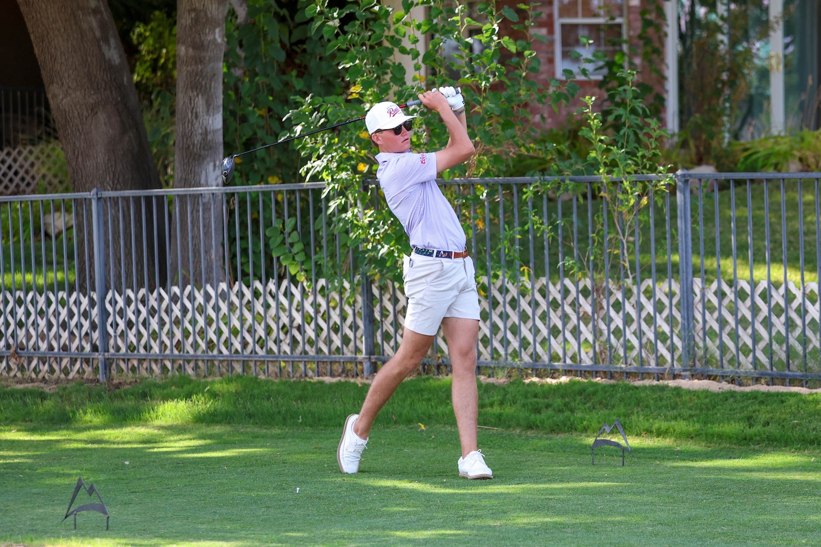 A person in a light polo shirt, white shorts, and cap mid-swing with a golf club on a grassy backyard range, fence and trees behind.