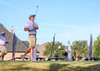 20260814-CoorsTOC-Day2-003 | Press Pass Sports Golfer in a purple polo and beige shorts preparing to swing on an elevated tee during a sunny golf event, with blue sky and flags in the background.