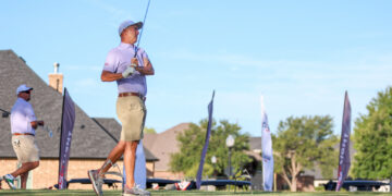 Golfer in a purple polo and beige shorts preparing to swing on an elevated tee during a sunny golf event, with blue sky and flags in the background.