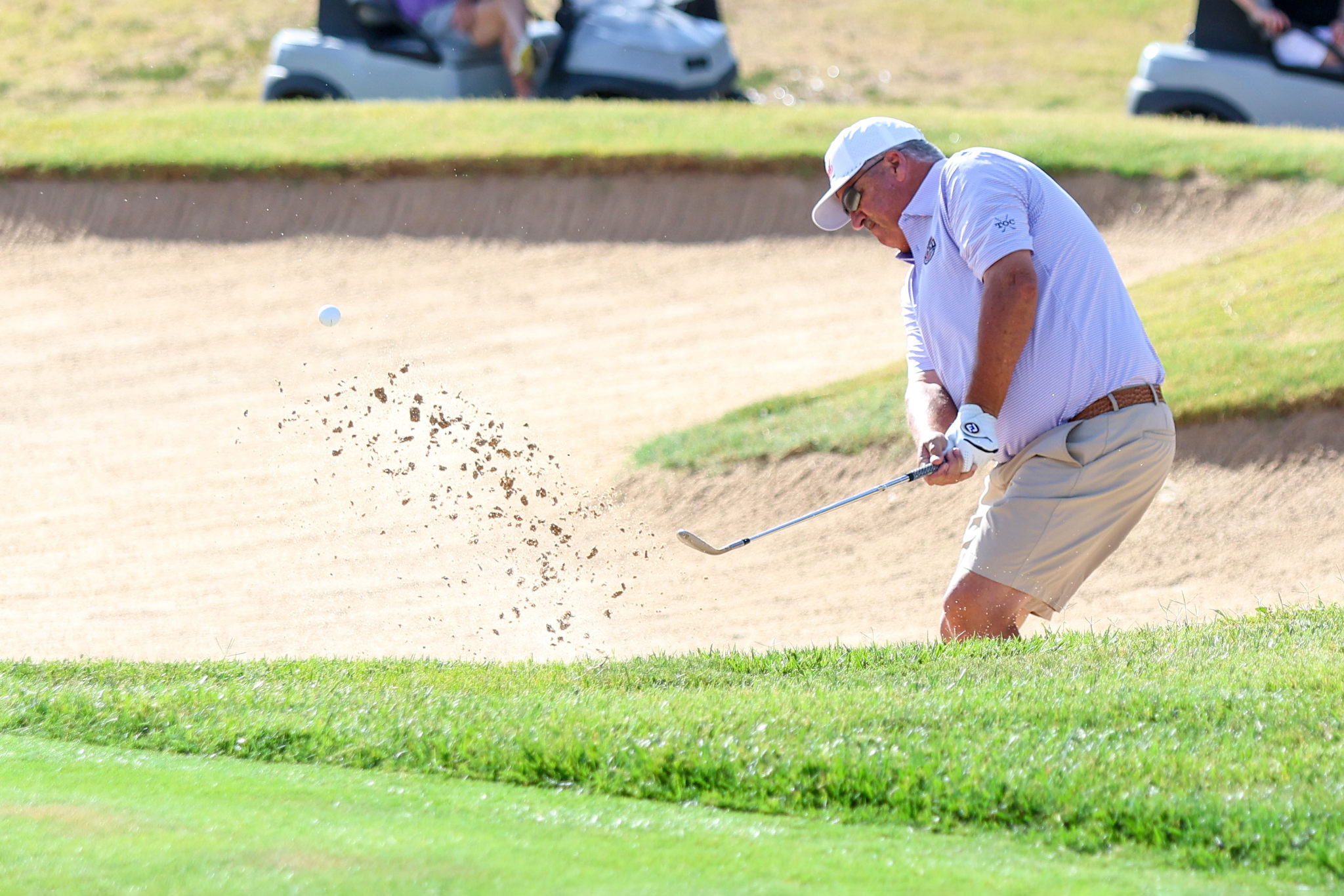 Golfer in a white cap and striped polo hits a bunker shot, sand spraying as the ball rises.