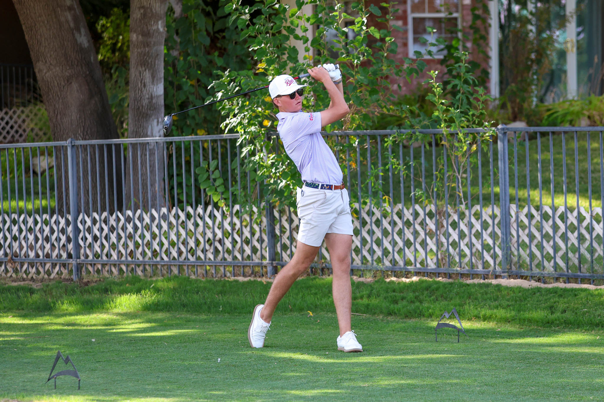 Man in white cap and sunglasses swinging a golf club in a backyard, mid-backswing with a metal fence and greenery in the background.