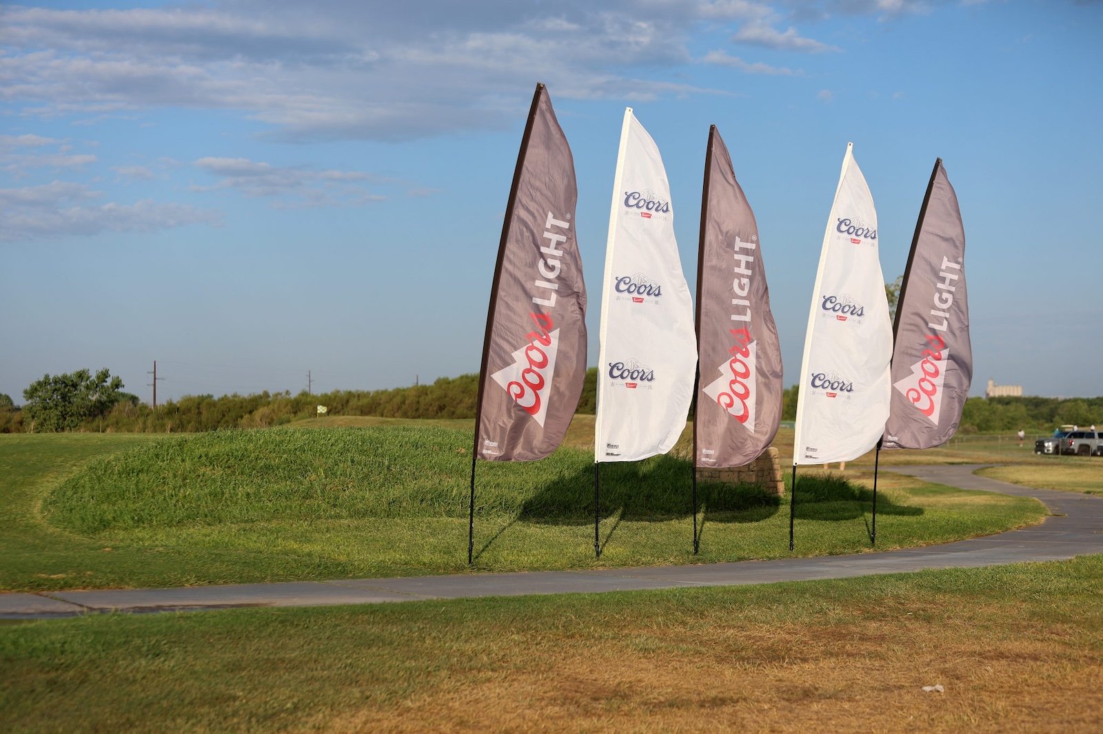 Row of Coors Light banners along a curved grassy golf course path under a blue sky.